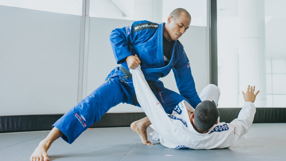Two athletes practicing Brazilian Jiu-Jitsu on a mat, one wearing a blue gi performing a technique while the other is on the ground.