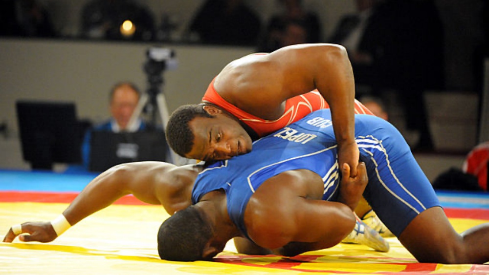 Two wrestlers engaged in a match, one in a red uniform and the other in a blue uniform, demonstrating a grappling move on a colorful wrestling mat.